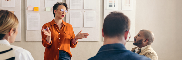 A business professional stands and speaks to a group of colleagues.